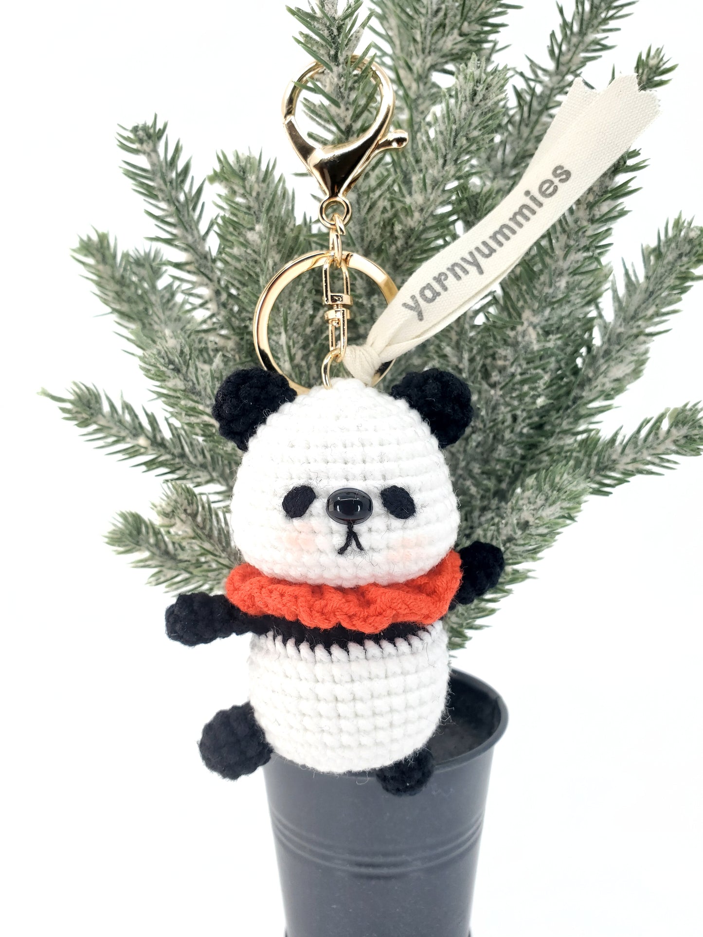 A small, plush keychain shaped like a panda with a red scarf, made from white cotton yarn, displayed on a green plant.