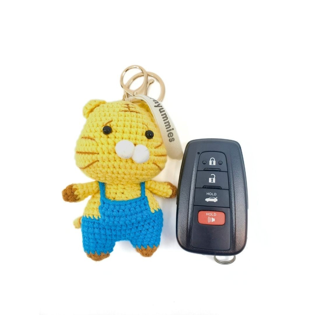 Crocheted cat keychain with blue overalls next to a car key fob on a white background