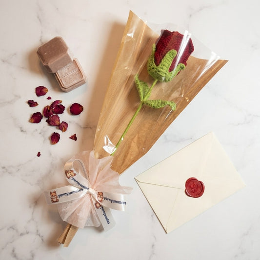 Crocheted rose in clear packaging with a small box and envelope on a marble surface