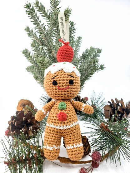 Handmade amigurumi Gingerbread Man with stitched eyes, smiling, wearing a Santa hat, displayed among Christmas decorations.