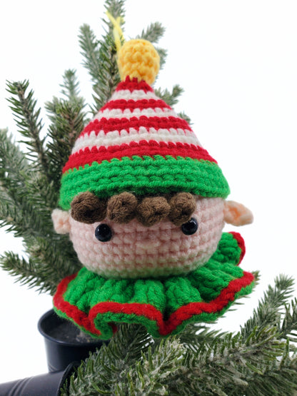 A handmade crochet elf head decoration with vibrant Christmas green and red colors, featuring a striped hat, displayed on a pine tree.