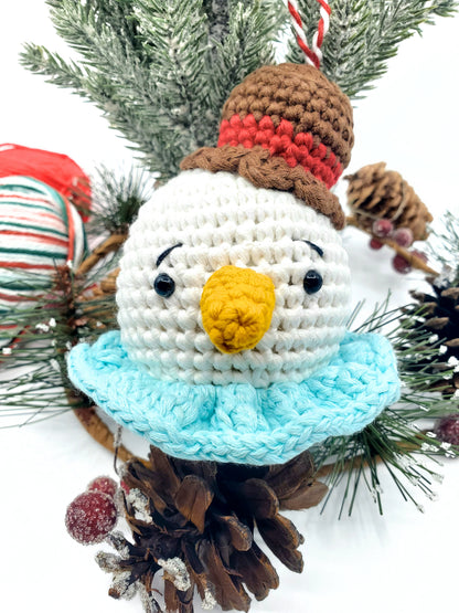 A crochet snowman head ornament with a red scarf, white hat, and a yellow nose, displayed among Christmas decorations.