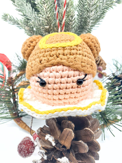 Handcrafted cotton yarn angel head ornament with a rattle, featuring brown hair, a yellow halo, and a white and pink robe, displayed against a festive Christmas tree background.
