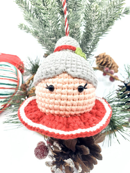 A crochet decoration resembling Mrs. Claus' head with a grey hat and red collar, displayed among Christmas tree decorations.