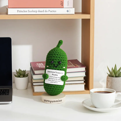 Green crocheted pickle plush with a label on a desk with books and a laptop.