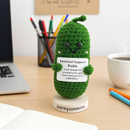 Green crochet pickle with a sign on a desk next to a laptop and coffee cup