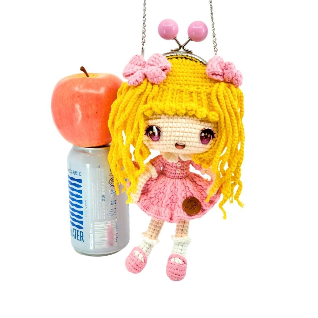 Crocheted doll with pink dress and yellow hair next to a water bottle and apple on a white background