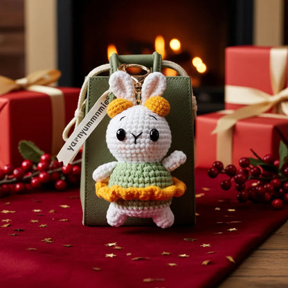 Small crocheted rabbit toy with a green dress on a gift box against a festive background with red berries and wrapped gifts.