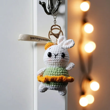 Crocheted bunny keychain with green dress and orange bow, hanging on a hook with 'yarnyummy' label.