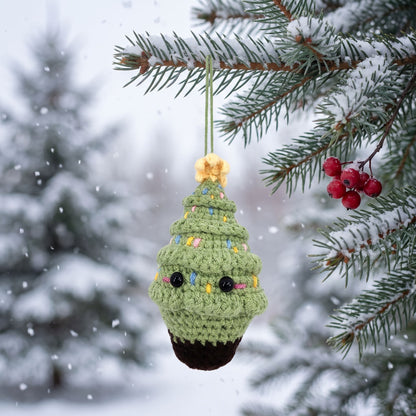 Handmade crochet Christmas tree ornament by YarnYummies hanging from a snowy pine branch outdoors during a snowfall. The ornament is light green with colorful yarn accents, black bead eyes, and a dark brown base, with a small gold star on top.