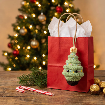 A Handmade crochet Christmas tree ornament by YarnYummies hanging from a red gift bag filled with white tissue paper, set on a wooden table with a blurred Christmas tree, candy canes, and gold chocolates in the background.