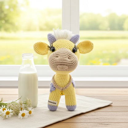 Crocheted yellow cow plushie next to a bottle of milk on a wooden surface with a window in the background.
