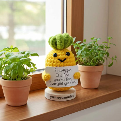 Yellow crocheted pineapple plush decor with a sign on a wooden surface with plants