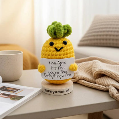 Crocheted pineapple plush with a face and text on a wooden stand, placed on a table.