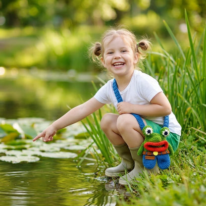 Child in green shorts with a frog designed crossbody bag, sitting by a pond in a natural setting.