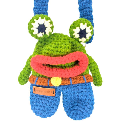 Crocheted frog character with a blue pants and strap on a white background