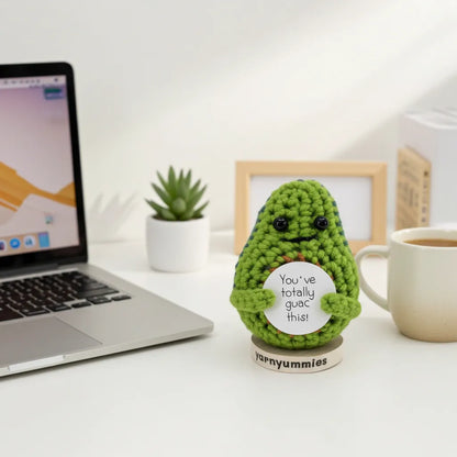 Green crocheted guacamole shaped plush with a message on a desk next to a laptop and a cup of coffee
