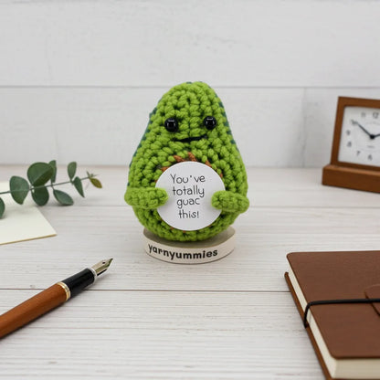 Crocheted avocado plush with a sign on a wooden surface, surrounded by stationery items.