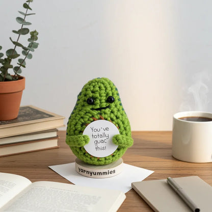 Green crocheted avocado plush decor holding a sign on a desk with books and a coffee cup.