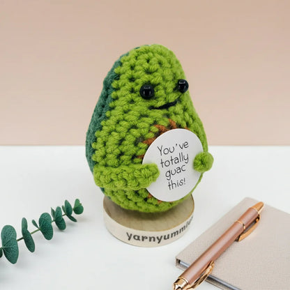 Crocheted avocado plush holding a sign with text, placed on a white surface with a pen and plant.