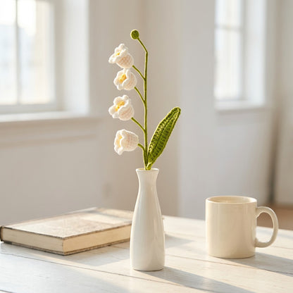 White vase with crocheted white may bells flowers on a table next to a book and a mug