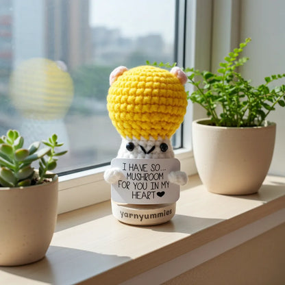 Crocheted mushroom character plush  with a yellow head on a windowsill with plants.