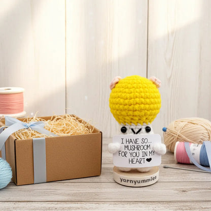 Yarnymummies crocheted mushroom character plush with a message on a wooden surface with yarn and a box in the background