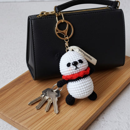 White crochet panda keychain with red scarf attached to keys, placed on a wooden surface with a black handbag in the background.