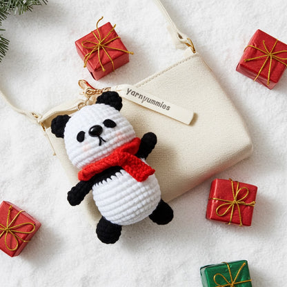 Crocheted panda keychain with red scarf on a white surface with small gift boxes.
