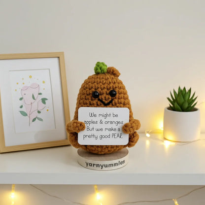 Crocheted pear character holding a sign with text on a shelf with decorative items.