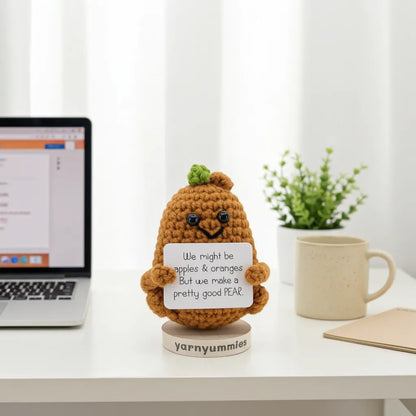 Crocheted pear-shaped character with a message on a desk next to a laptop and mug.