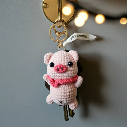 Crocheted pig keychain with pink scarf on a key against a neutral background