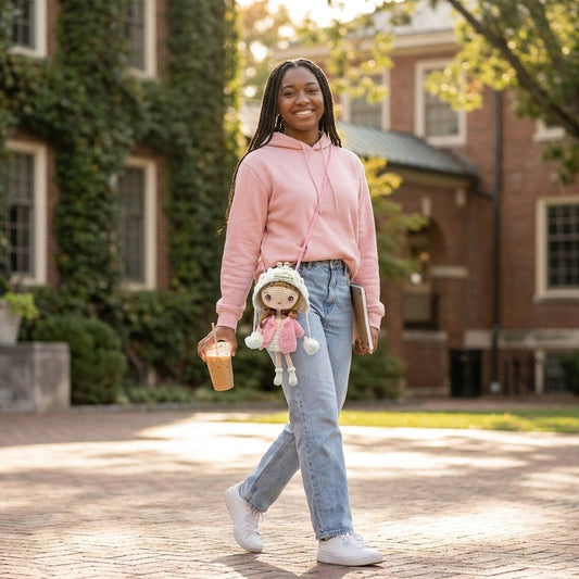 Person in pink hoodie and jeans holding a coffee cup and doll, standing on a brick path with a building in the background.