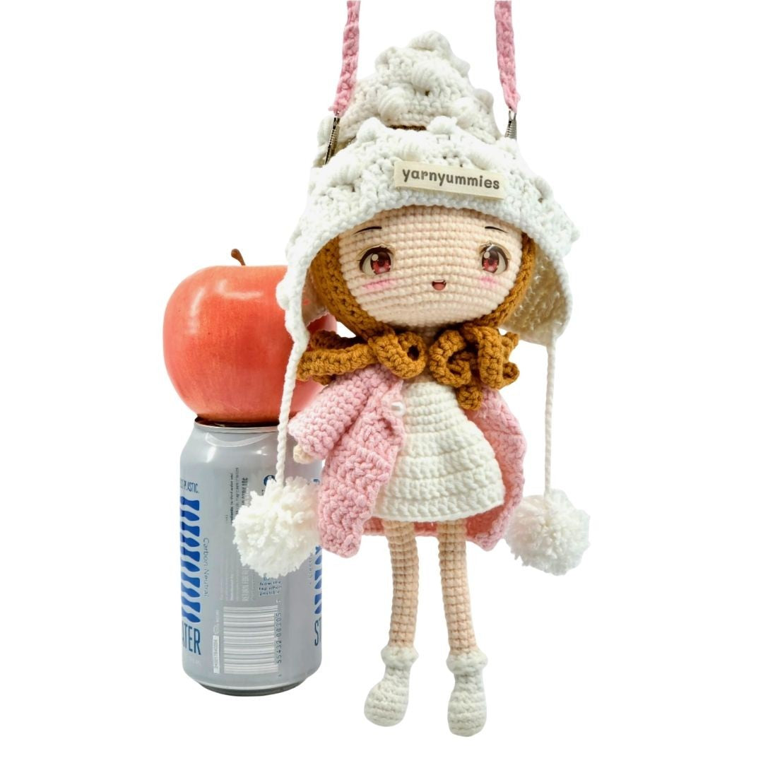 Crocheted doll with a pink dress and white hat, standing next to an apple and a can on a white background.