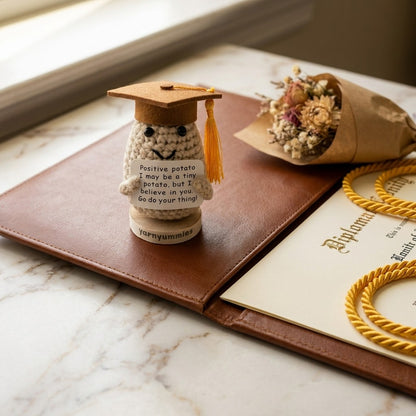 Small crocheted positive potato character with a graduation cap on a marble surface with a diploma and flowers.