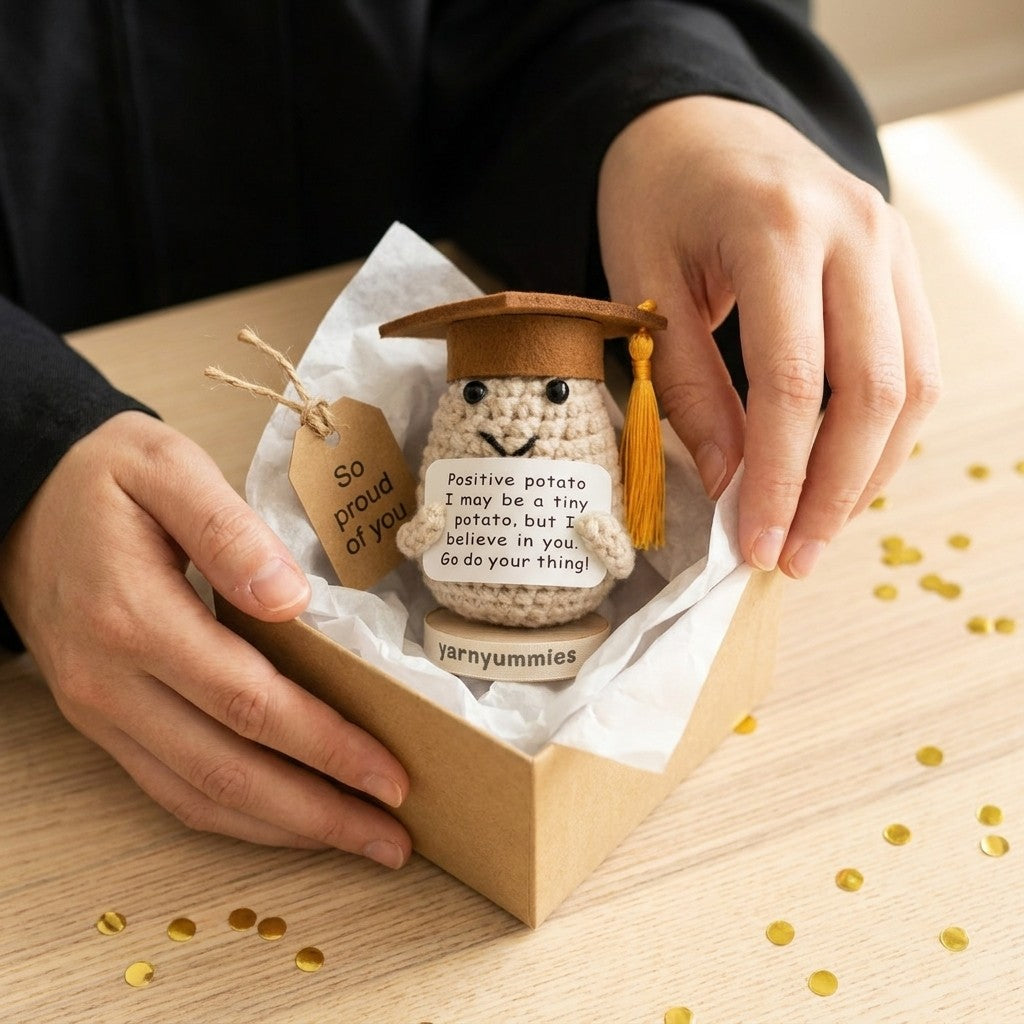 Small crocheted positive potato character plush with a graduation cap in a box, held by hands on a wooden surface.