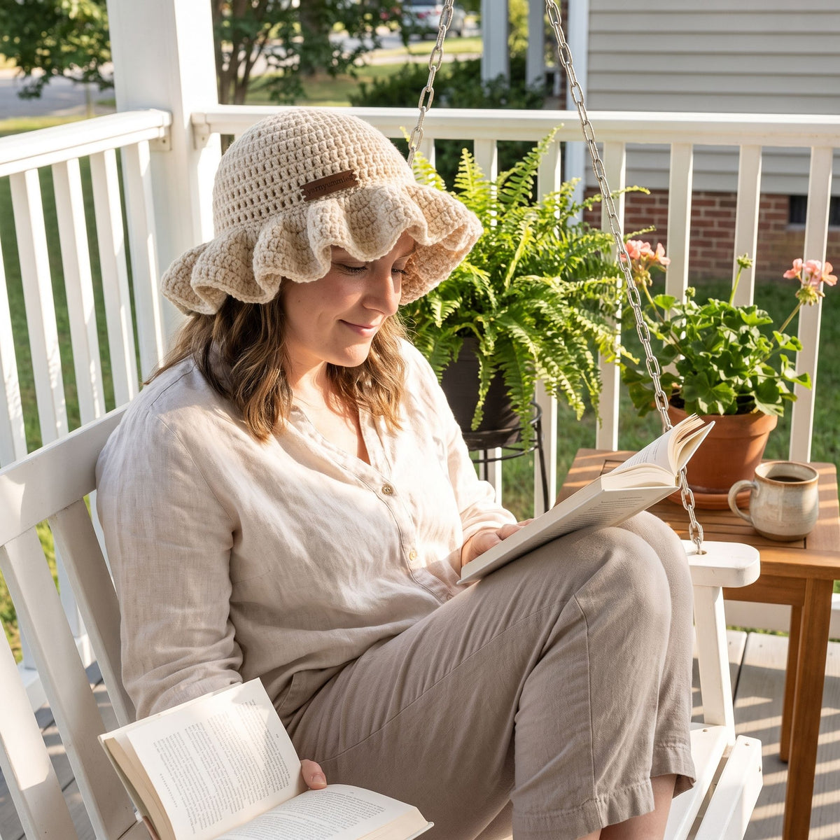 Woman reading a book on a front porch swing wearing a cream handmade crochet ruffled brim bucket hat by YarnYummies