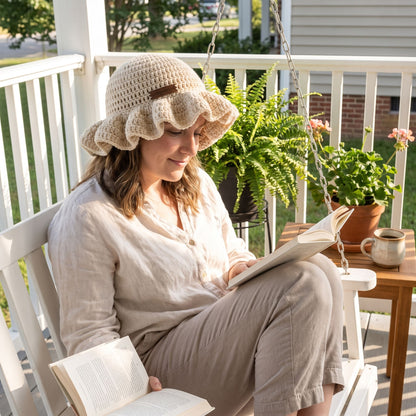 Woman reading a book on a front porch swing wearing a cream handmade crochet ruffled brim bucket hat by YarnYummies