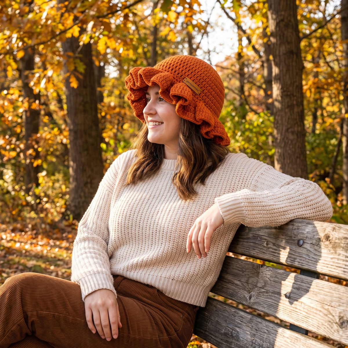 Woman smiling on a park bench wearing an orange handmade crochet ruffled brim bucket hat by YarnYummies in an autumn forest setting