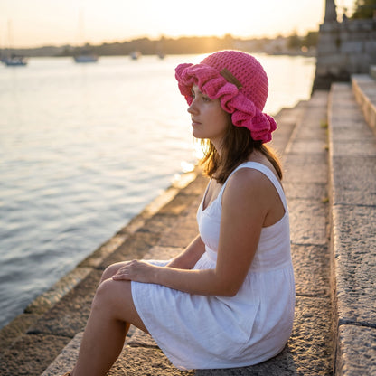 Woman sitting on a waterfront at sunset wearing a pink handmade crochet ruffled brim bucket hat by YarnYummies