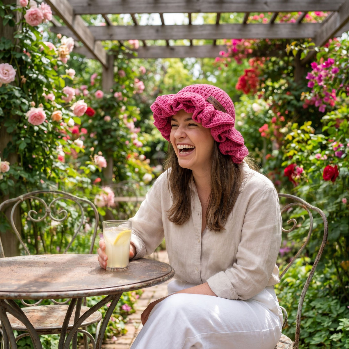 Woman laughing at a garden table wearing a pink handmade crochet ruffled brim bucket hat by YarnYummies surrounded by blooming roses