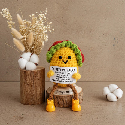 Amigurumi taco plush toy with a message on a wooden surface with decorative elements.
