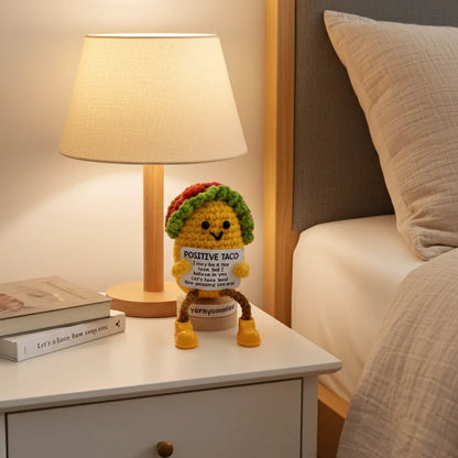 Crocheted plush toy resembling a taco with a sign on a nightstand next to a lamp and books.