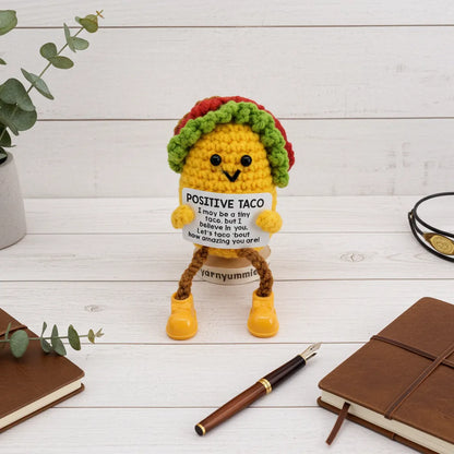 Crocheted taco character holding a sign with text on a wooden surface with books and a pen.