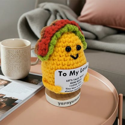 Crocheted taco plush toy with a 'To My Love' label on a table with a mug and magazine.