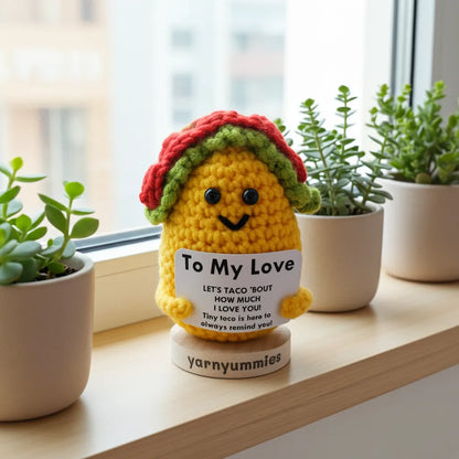 Crocheted taco plush toy holding a card with text, placed on a windowsill with plants.