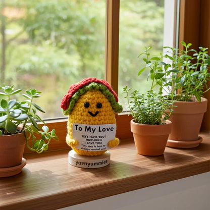 Crocheted taco-shaped figure with a message card on a windowsill with plants