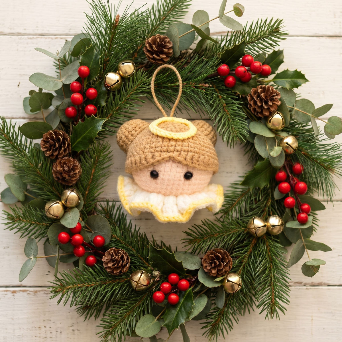 A handmade crocheted angel head ornament with a yellow halo by YarnYummies, paced at the center of a green eucalyptus and pine wreath decorated with red berries, pinecones, and small gold bells on a white wood table.