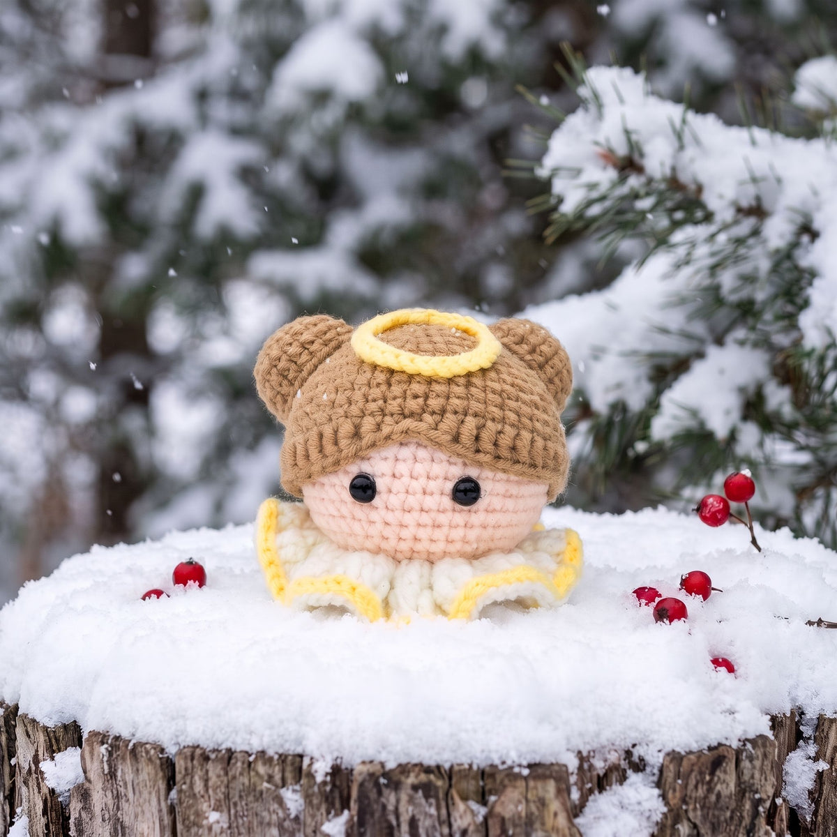 A Handmade crochet angel head ornament by YarnYummies front view with a tan hat and halo sits on a snowy log outdoors, with snow-covered evergreen branches and red berries in the background.