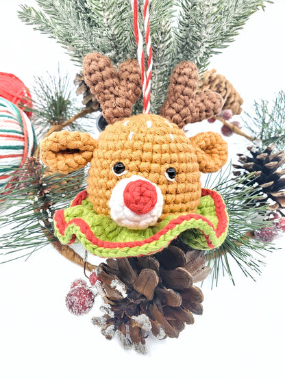 A crochet reindeer head with rattle, featuring a red nose and green collar, placed among Christmas tree decorations.
